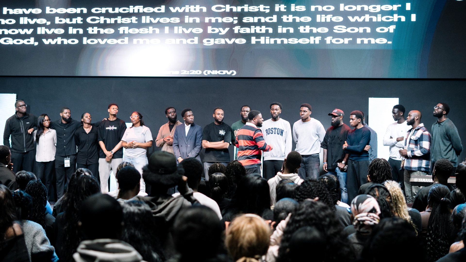 A wide stage with Galatians 2:20 projected; a line of young men stands across the front as the congregation looks on.