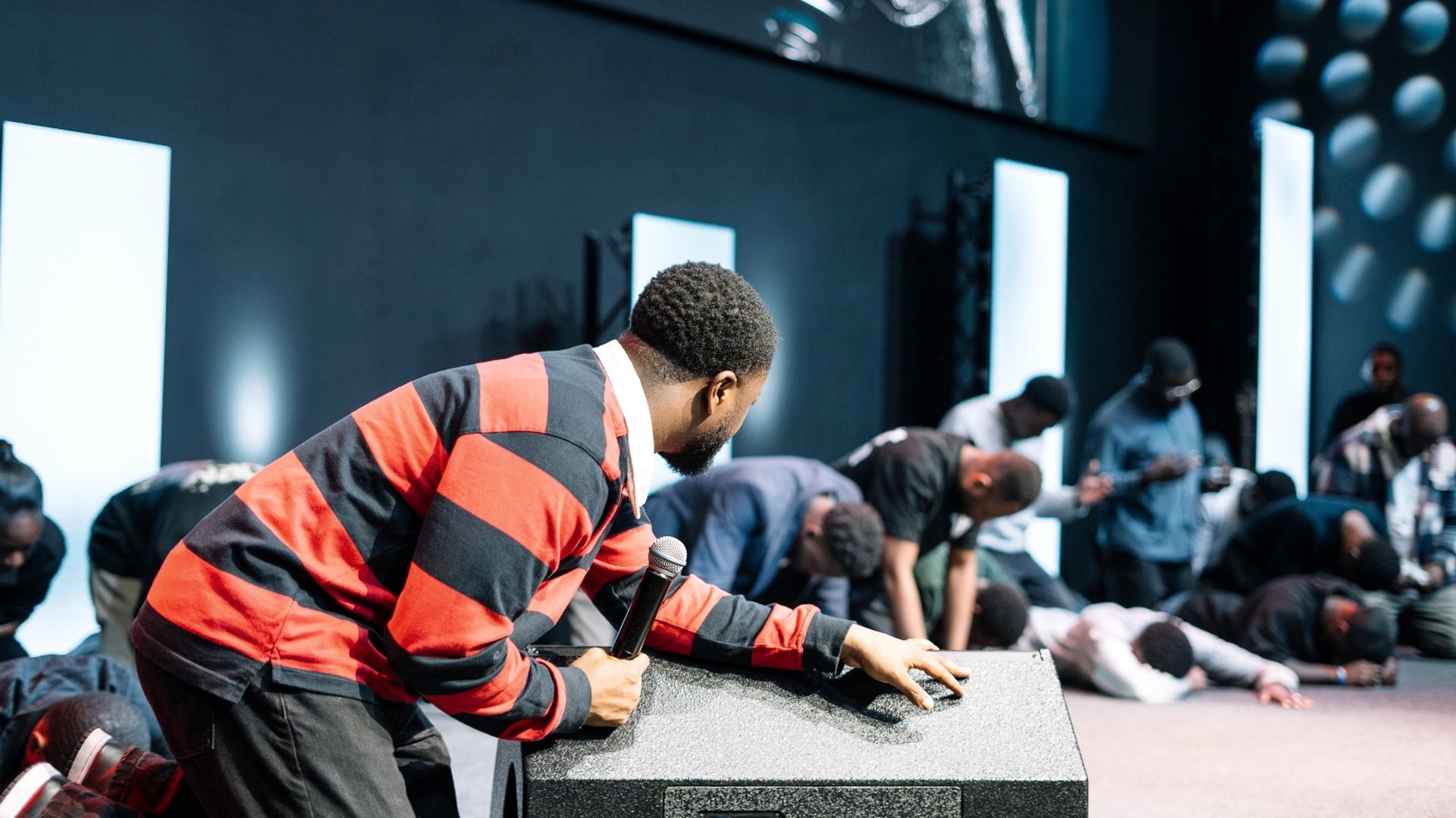 A pastor leaning on a monitor as the congregation kneels in prayer.